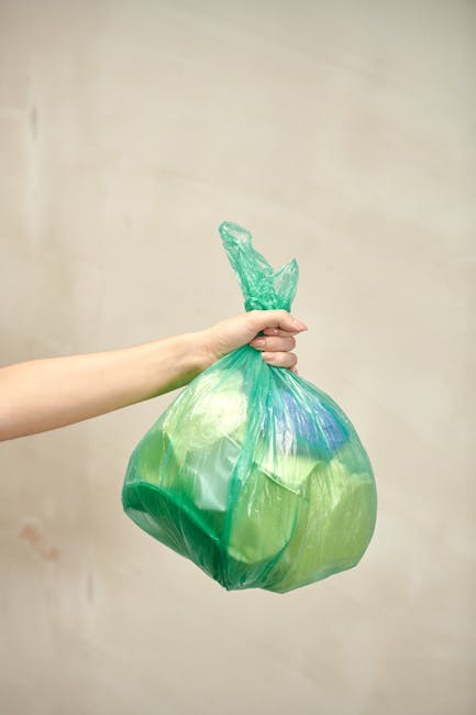 A person's hand holding a green plastic bag filled with household waste, including a crumpled plastic bottle at the top. The background features a plain, light-colored wall. The image relates to the disposal of bulky waste such as sofas and mattresses, which are typically handled during house removals or furniture transport. The scene suggests waste collection or clearance as part of a home relocation process, highlighting the importance of proper waste disposal rules in Greenford. The lighting is neutral and even, emphasizing the details of the plastic bag and its contents, consistent with professional removal services like Man with Van Greenford that assist with packing, moving, and waste disposal.