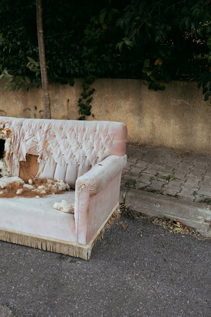A discarded, worn-out pink velvet sofa with built-in tufted cushioning and floral detailing on the left armrest, positioned outdoors on a paved area next to a concrete wall overgrown with green foliage. The sofa has torn fabric, exposing foam padding and stuffing inside, indicating it is no longer suitable for use. It is situated partially on the pavement and partially on the uneven ground, with no surrounding furniture or items nearby. The scene is illuminated by natural daylight, and the background features a weathered wall with some greenery creeping over the top, suggesting a residential or back garden area. This image illustrates typical furniture removal scenarios encountered during home relocation or clearance services, such as those provided by Man with Van Greenford, which handle the disposal and transportation of bulky waste items like sofas.