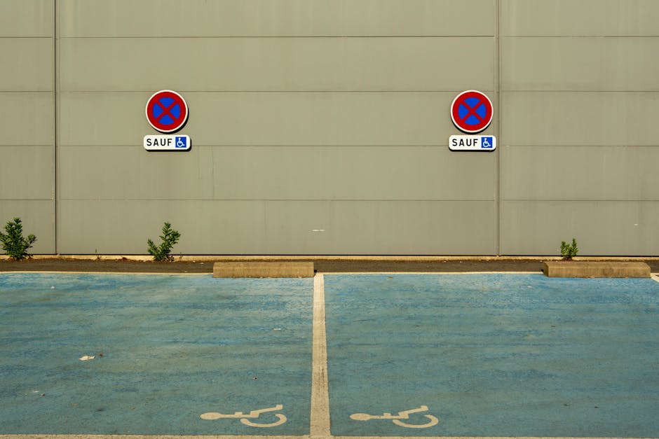 A photograph of a parking area with two designated accessible parking spaces marked by blue painted lines and wheelchair symbols on the ground. Each space is separated by a white line and features a concrete wheel stop at the end closest to a plain beige wall. Two prominent parking restriction signs are mounted on the wall above the spaces, each displaying a red circle with a blue background and a red slash indicating no parking, with a small white label underneath reading 'SAUF' and a blue symbol of a wheelchair, indicating exceptions for disabled parking. The space on the right is partially blocked by a small weed growing beside the concrete wheel stop, while the left space is clear. Behind the parking spaces, the beige wall has horizontal lines and appears smooth, with diffuse natural lighting illuminating the scene evenly. The setting suggests a commercial or residential parking lot, with a clean pavement and minimal debris, and occasional small plants growing at the base of the wall, providing a neutral backdrop for the parking restrictions relevant in the context of house removals and furniture transport logistics conducted by Man with Van Greenford.