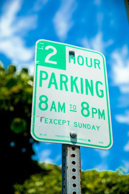 A close-up view of a green and white rectangular parking restriction sign mounted on a metal pole, indicating a two-hour parking limit from 8 a.m. to 8 p.m., Monday to Saturday, with a note that parking is not permitted on Sundays. The sign features bold green text and a green border, with a small black screw fastening it to the pole. In the background, there are blurred green foliage and a bright blue sky with some scattered clouds. The scene is outdoors, suggesting an urban or residential area where house removals or furniture transport services by Man with Van Greenford operate, requiring careful parking enforcement and adherence to local parking regulations during home relocation or moving projects.