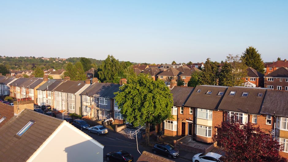 Aerial view of a residential street in Greenford showing a row of terraced houses with pitched roofs, some with skylights, and front yards with parked cars. A large, leafy green tree is centrally positioned along the pavement, casting a shadow on the road. The houses feature a mix of brick and weatherboard exteriors, with some windows open or featuring bay windows. The street appears calm with no visible moving activity; however, the scene suggests a typical environment where home relocation or furniture transport services by Man with Van Greenford might take place, supported by the spacious frontage suitable for loading and unloading goods during house removals or furniture transport. The clear sky and daylight provide good lighting, highlighting the details of the properties, vehicles, and greenery. This setting visually aligns with property relocation tasks including packing, moving logistics, and loading processes.