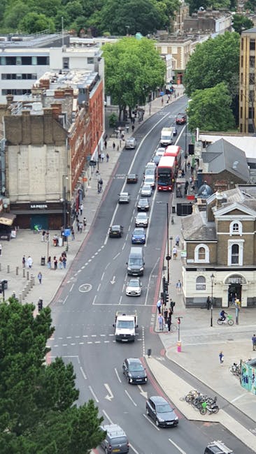 An aerial view of a busy residential street in Greenford, featuring a line of parked vehicles along the curb, with several moving cars traveling in both directions. On the right side, there are a variety of buildings including a small, white, single-story house with a pitched roof and arched windows, and a larger brick building with multiple levels. The street is bordered by tall, leafy green trees providing shade, and pedestrians are visible walking along the sidewalks. In the foreground, bicycles are parked near a corner, and a cyclist is riding on the pavement. The environment is well-lit with natural daylight, and the scene captures typical urban activity during daytime, relevant to house removals, local transportations, or furniture transport involving [COMPANY_NAME] in the context of moving and packing processes in Greenford.