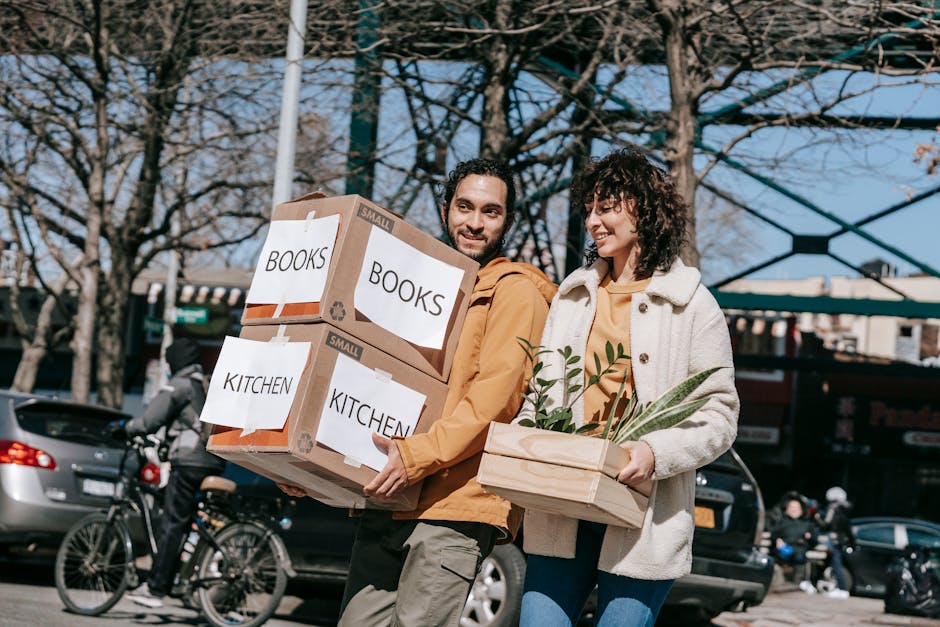 A man and a woman are outdoors during daytime, engaged in a house relocation process, as part of packing and moving services. The man, with dark hair and a beard, is wearing a yellow jacket and is holding two cardboard boxes labeled 'BOOKS' and 'KITCHEN'. The boxes are taped and reinforced with plastic wrap, indicating careful packing. The woman, with curly hair, is dressed in a light-colored jacket and is carrying a wooden crate containing green houseplants, illustrating the transfer of household items. They are standing on a sidewalk, with a background featuring leafless trees, parked cars, bicycles, and a building structure, suggesting an urban setting. The scene is well-lit, capturing the active loading process associated with home removals managed by Man with Van Greenford. The image visually emphasizes organized packing, safe transport of household belongings, and the movement of furniture and items during a residential move.