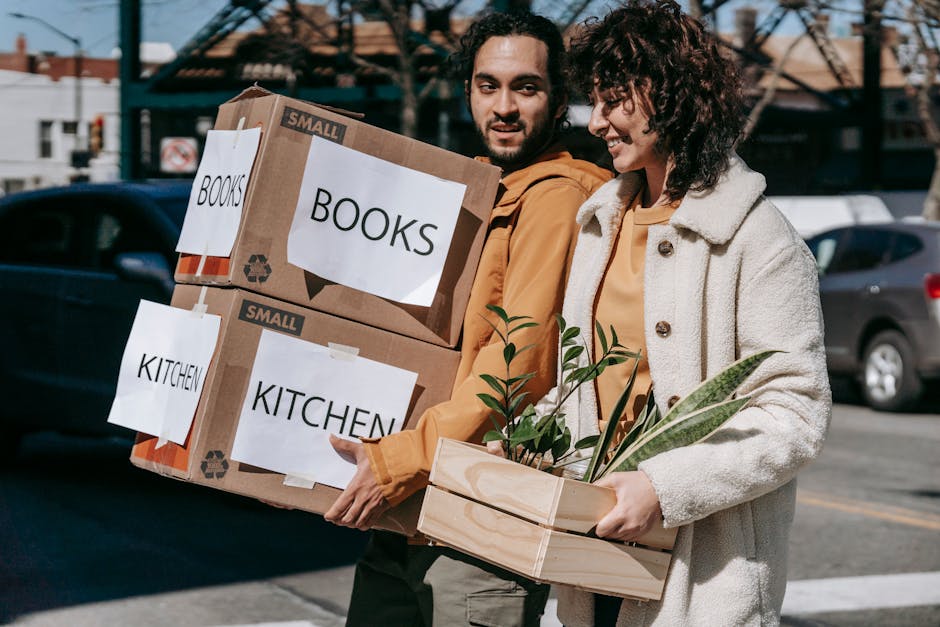 A man and woman engaged in house relocation activities outdoors beside a parking area, with several cardboard boxes labeled 'BOOKS' and 'KITCHEN' being carried. The man, with dark curly hair and a beard, is holding two small cardboard boxes stacked one on top of the other, while the woman, with curly brown hair, is holding a wooden crate containing potted plants. The boxes are sealed with packing tape and paper labels indicating their contents, designed to assist in organized packing and furniture transport during a house move. Behind them, a dark vehicle is parked, with an urban backdrop featuring buildings and a clear sky. The scene is lit naturally, suggesting daytime, and depicts a dynamic loading process which is typical during home relocation using professional removal services. This image exemplifies packing and moving logistics as part of a comprehensive house removal, with a focus on careful handling of household items and plants, aligning with the services offered by Man with Van Greenford.