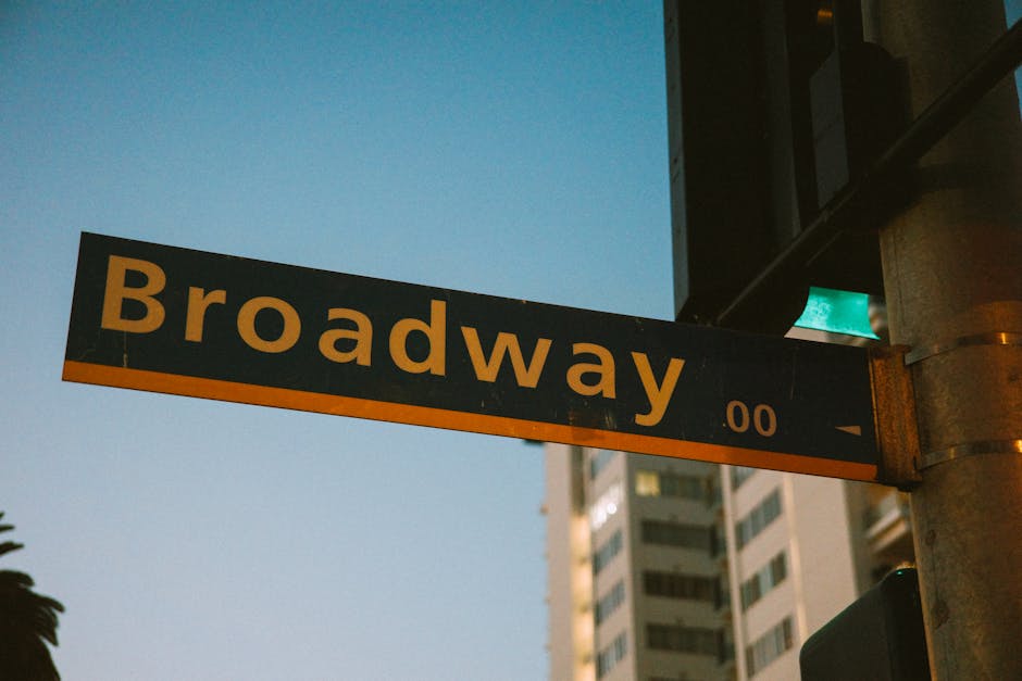 A man and a woman are outdoors during daytime, engaged in a house relocation process, as part of packing and moving services. The man, with dark hair and a beard, is wearing a yellow jacket and is holding two cardboard boxes labeled 'BOOKS' and 'KITCHEN'. The boxes are taped and reinforced with plastic wrap, indicating careful packing. The woman, with curly hair, is dressed in a light-colored jacket and is carrying a wooden crate containing green houseplants, illustrating the transfer of household items. They are standing on a sidewalk, with a background featuring leafless trees, parked cars, bicycles, and a building structure, suggesting an urban setting. The scene is well-lit, capturing the active loading process associated with home removals managed by Man with Van Greenford. The image visually emphasizes organized packing, safe transport of household belongings, and the movement of furniture and items during a residential move.