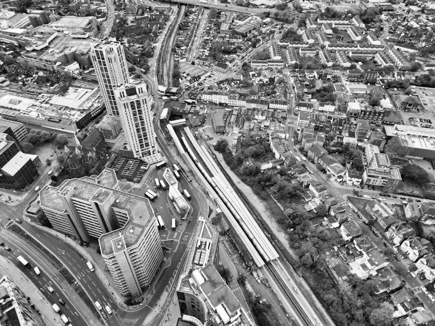 Black and white aerial photograph of a dense urban area featuring a mix of high-rise and mid-rise buildings, with a prominent railway station platform visible near the centre. Multiple buses and cars are seen on surrounding roads, indicating busy traffic flow. A railway line runs parallel to a street lined with commercial and residential structures, and the platform appears to be in use with some objects possibly related to moving or transportation logistics. The presence of a large wooden crate, cardboard boxes, and wrapped furniture around the station suggests ongoing home relocation or furniture transport activities. These items are situated close to the platform and adjacent pavement, with some partially inside a vehicle or trolley for loading. Overall, the scene captures a busy city environment engaged in packing, loading, and moving processes, characteristic of professional removals services like those offered by Man with Van Greenford as seen in the context of house removals and moving logistics.