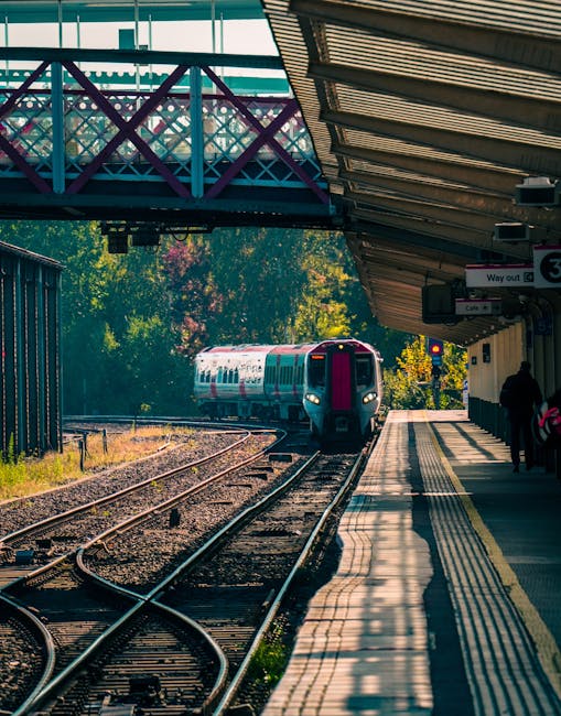 A modern passenger train arriving at Greenford station on a sunny day, with the platform on the right side featuring tactile paving strips along the edge and a small group of passengers waiting under an overhanging shelter. The station canopy extends over the platform, providing shade for waiting travelers, while a pedestrian footbridge painted in purple and teal crosses above the railway tracks. The surrounding area includes lush green trees, contributing to a calm, suburban atmosphere. The railway tracks are visible in the foreground, leading towards the approaching train, which has a sleek design with a white and purple exterior. The scene captures the typical setting of a UK suburban train station involved in daily relocation and commuting activities, with an emphasis on transportation infrastructure and the environment supporting house removals and furniture transport logistics.