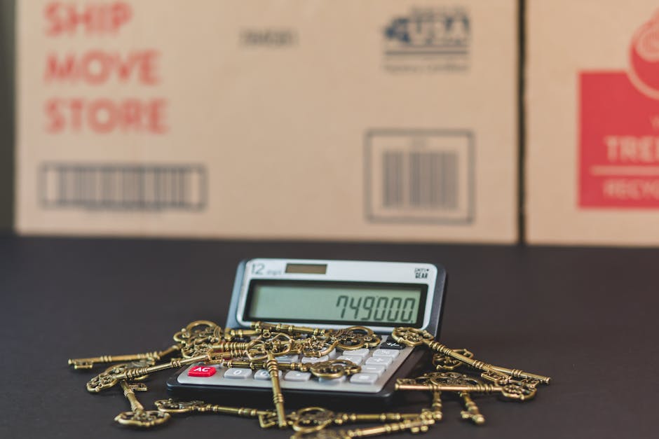 A digital calculator displaying the number 749,800 is placed on a black surface, with several gold-colored keys and key-shaped objects scattered across it. In the background, there are partially visible cardboard boxes with red and black printed labels, including words like 'Ship,' 'Move,' and 'Store,' indicating moving or packing supplies. The boxes are positioned against a plain, light-colored wall. The scene suggests a focus on the costs associated with home relocation or furniture transport, with the presence of packing materials and the calculator symbolizing moving expenses. Man with Van Greenford's removals service is subtly referenced through the context, highlighting the logistics and costing aspect of house removals.