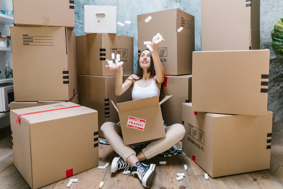 A woman sitting inside a room surrounded by multiple large cardboard moving boxes, some sealed with red tape and others open, with packing foam peanuts scattered around her. She is wearing casual clothing, including a white tank top and sneakers, and is holding packing foam peanuts in her hands while looking up. The boxes are stacked against a wall with a rough, textured surface and a small green plant is visible in the corner. The floor is wooden, and a doorway is partially visible on the left side of the image. This scene depicts the packing and home relocation process managed by [COMPANY_NAME], illustrating the careful handling of boxes and packing materials during furniture transport or moving services.