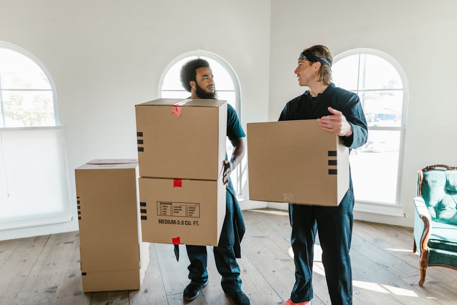 Two men engaged in a home relocation process inside a well-lit room with light-colored walls and hardwood flooring. One man with curly hair and a beard is holding a large cardboard box, while the other man with straight hair and wearing a headband is also carrying a box. Several additional cardboard boxes of varying sizes, some labeled with weight and volume information, are positioned on the floor nearby. The room features large arched windows allowing natural light to illuminate the space, and there is a green upholstered armchair with wooden accents against the wall. Behind the men, a doorway is visible, and the overall setting suggests a furniture transport or packing and moving activity, typical of a professional removal service such as Man with Van Greenford, preparing for or executing a house move.