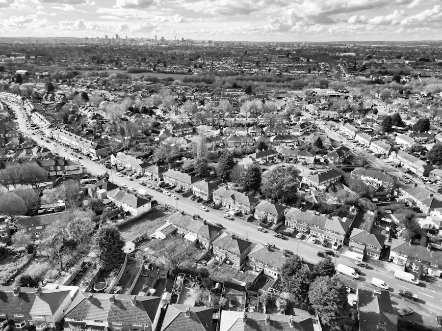Black and white aerial photograph showing a suburban residential area with a main street lined with terraced and semi-detached houses, some with driveways and small gardens. Several parked cars are visible along the street and in driveways. In the foreground, rooftops of individual houses with chimneys and a mix of tiled and flat roofs can be seen, some surrounded by trees and garden fences. The street appears to be in the process of a home relocation or furniture transport, with a van parked on the pavement near the houses, likely associated with Man with Van Greenford’s removals services. The background extends to an open landscape with fields and distant city skyline, under a partly cloudy sky, indicating daytime. The scene captures the loading process typical of home removals, including moving boxes, furniture, and packing materials possibly prepared for a same-day move in Greenford, aligned with the page's content about last-minute moves.