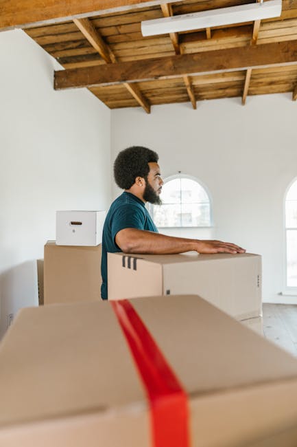 Two men engaged in a home relocation process inside a well-lit room with light-colored walls and hardwood flooring. One man with curly hair and a beard is holding a large cardboard box, while the other man with straight hair and wearing a headband is also carrying a box. Several additional cardboard boxes of varying sizes, some labeled with weight and volume information, are positioned on the floor nearby. The room features large arched windows allowing natural light to illuminate the space, and there is a green upholstered armchair with wooden accents against the wall. Behind the men, a doorway is visible, and the overall setting suggests a furniture transport or packing and moving activity, typical of a professional removal service such as Man with Van Greenford, preparing for or executing a house move.