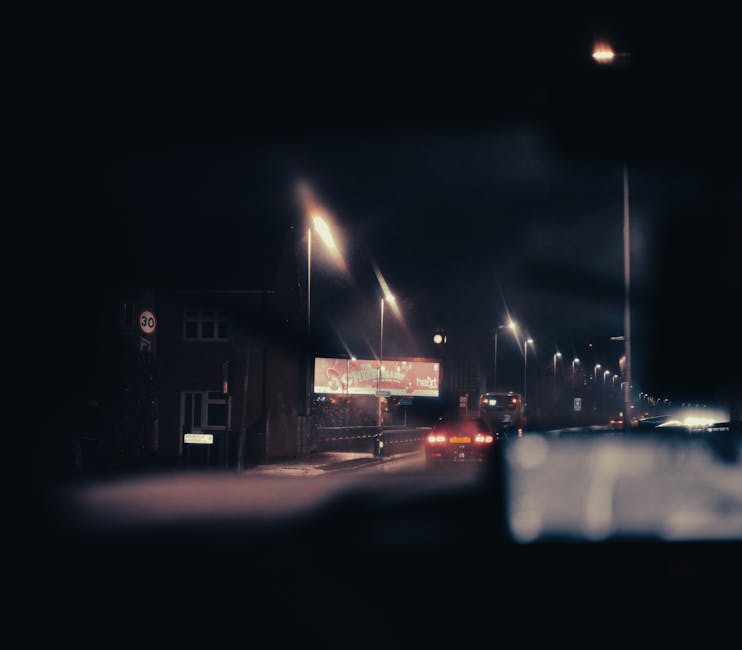 A nighttime view from inside a vehicle showing a narrow residential street in Greenford, UB6, illuminated by streetlights. Visible on the pavement are parked cars, and the roadside features a building with a brightly lit advertising billboard. The street is dimly lit, with glow from the streetlights highlighting the road and parked vehicles, while the dark sky is visible overhead. This environment may relate to house removals or transport activities involving Man with Van Greenford, particularly in navigating tight access points during a home relocation or furniture transport process. The scene emphasizes the confined urban street setting typical for local moves, with focus on the limited space for vehicle maneuvering and careful loading or unloading of furniture and packing materials near the property address specified on the page about house moves on Oldfield Lane.
