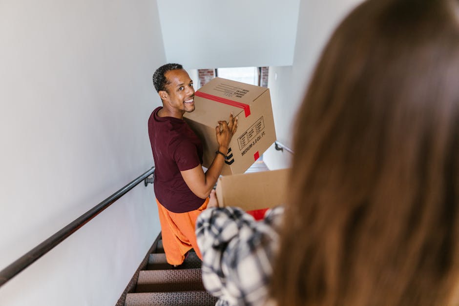 A man with dark skin, short curly hair, wearing a maroon t-shirt and orange trousers, is positioned on a staircase inside a residential property, smiling while holding a medium-sized cardboard box with red tape and printed labels, indicative of home relocation and packing. The staircase has a black metal handrail on the left side, and the wall is painted white. In the foreground, the back of a woman with long brown hair and a plaid shirt is partly visible, holding a cardboard box, suggesting active packing and loading processes carried out by Man with Van Greenford, a professional removals company. Natural light filters through a window at the top of the stairs, illuminating the scene in a neutral, functional manner, emphasizing the careful handling and transportation of household items involved in furniture transport and moving logistics.