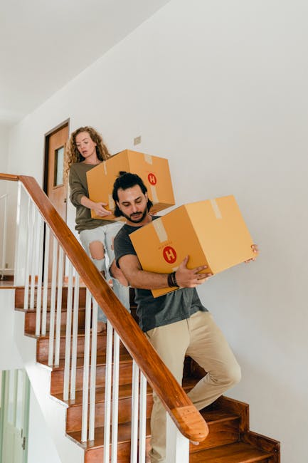 Two movers from Man with Van Greenford, dressed in casual clothing, are carrying large cardboard boxes with red labels down a wooden staircase inside a residential property. One mover, positioned on the lower part of the staircase, is stepping carefully while holding a box close to his chest, with a focused expression. The second mover, further up the stairs, is also gripping a box and guiding it down alongside him. The staircase features a polished wooden handrail and white-painted railing spindles, with a neutral-toned wall and a wooden door visible in the background. The scene captures a home relocation process involving the careful handling and transportation of boxed belongings, reflecting professional packing and moving activities by Man with Van Greenford. Proper lighting ensures details of the boxes, clothing, and staircase are clearly visible, providing an accurate visual representation of furniture transport within a residential setting.