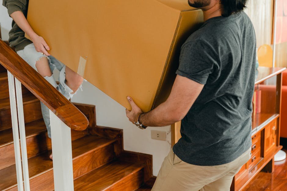 A man with dark skin, short curly hair, wearing a maroon t-shirt and orange trousers, is positioned on a staircase inside a residential property, smiling while holding a medium-sized cardboard box with red tape and printed labels, indicative of home relocation and packing. The staircase has a black metal handrail on the left side, and the wall is painted white. In the foreground, the back of a woman with long brown hair and a plaid shirt is partly visible, holding a cardboard box, suggesting active packing and loading processes carried out by Man with Van Greenford, a professional removals company. Natural light filters through a window at the top of the stairs, illuminating the scene in a neutral, functional manner, emphasizing the careful handling and transportation of household items involved in furniture transport and moving logistics.