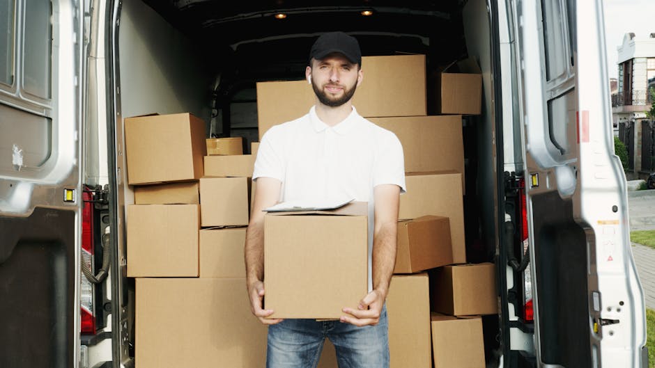 A man wearing a black cap, white polo shirt, and jeans is standing at the rear of a large moving van, holding a cardboard box. The van's interior is filled with neatly stacked cardboard boxes of vario
