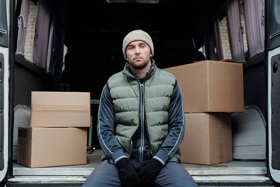 A man wearing a black cap, white polo shirt, and jeans is standing at the rear of a large moving van, holding a cardboard box. The van's interior is filled with neatly stacked cardboard boxes of vario